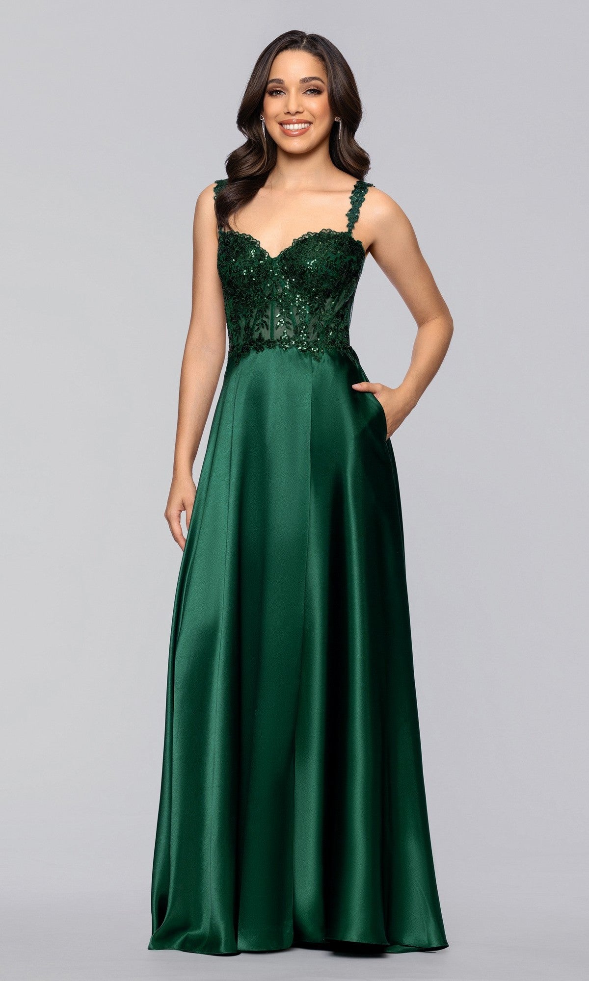 A woman stands against a light gray background wearing the Blondie Nites 6088BN long prom dress, featuring an a-line satin silhouette with a floor-length dark green skirt and a sleeveless, sequin-embellished bodice.